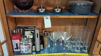 Photo showing multiple barware items including glassware, metal butter holders, and a ceramic casserole holder inside a wooden cabinet shelf.