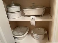 Overall view of five Corningware cookware pieces arranged on shelf: two white ceramic baking dishes in metal holders with glass lids, and two square Corningware dishes with blue floral pattern, all with lids.