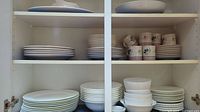 Two sets of stacked plates and bowls with cups and saucers on shelves inside a cabinet
