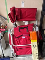 Three red folding camp chairs stacked and folded, showing the front and storage pockets of one chair along with a colorful box against a beige wall.