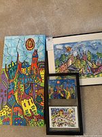 Three artworks displayed on carpet: left original colorful acrylic painting on canvas; top right large framed print with cityscape; bottom right smaller framed prints with bright abstract designs.