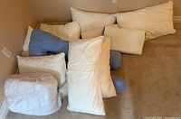Top view of assorted pillows laid on carpet including white regular pillows, blue pillows, and one pillow in plastic wrapping.