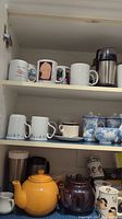 Photo showing shelves of various mugs, ceramic teapots in yellow and dark brown, blue floral patterned mugs and teacups, and stainless steel travel mug.