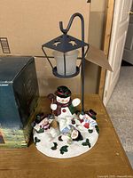 Photo of snowman Christmas candle holder featuring black metal lantern with frosted glass tealight holder and three snowmen figurines on a snowy base with holly leaves.