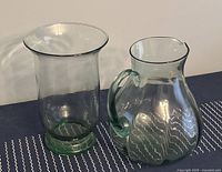 Tall green glass vase next to a green glass water pitcher, both with translucent green tint and smooth surfaces