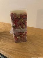 Close-up photo of large rectangular decorative candle filled with dried flowers and petals in red and green tones, wrapped with ribbon.