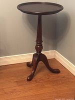 Small wooden round table with round top and three curved legs, dark finish, placed on hardwood floor against wall.