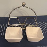 Two white ceramic square bowls with a silver-tone metal stand and spreader on blue cloth
