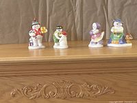 Four Royal Doulton snowman family figurines lined up on a wooden surface in front of a curtain background. Each figurine is uniquely posed with winter accessories.