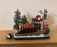 Christmas themed engine train with Santa figure on the train, small elf figure placing a stocking, pine tree in the background, and power adaptor on wooden surface.
