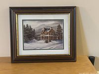 Framed print showing a large house covered in snow surrounded by trees, with brown wooden frame and white matting.
