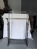 Photo showing an adjustable rolling clothing rack with a metal frame, single horizontal hanging bar, vertical supports, and wheeled base.