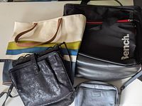 Four bags arranged on a flat surface: a striped canvas tote bag, a black and silver Bench yoga bag, a small black metallic crossbody purse, and a gray pebbled faux leather handbag.