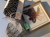 Photo of the moose watercolor painting framed in wood alongside two fabric plant hangers with rope.