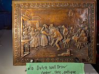 Front view of an antique Dutch copper decorative plate with detailed raised-relief scene showing several 17th-century styled figures in an interior, including men, women, children, a dog, furniture, and decorative elements.