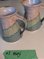 Two handmade ceramic mugs blurred in background, showing combined purple, green, and turquoise glaze with handles on the right side.
