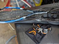 Squash racket with four squash balls placed on it and protective sports glasses with soft pouch underneath on a black surface.