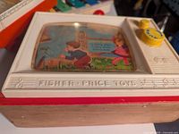Close-up of vintage Fisher-Price toy clock with white body, wooden base, and illustrated dial with children under plastic labeled FISHER-PRICE TOYS.