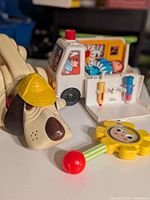 Beige dog rattle toy with yellow hat, alongside yellow flower-shaped rattle; visible wear and discoloration evident.