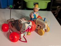Side view of the vintage Fisher-Price wooden dog pull toy featuring painted details, red wheels, and pull string, alongside the tractor toy with the farmer figure visible.