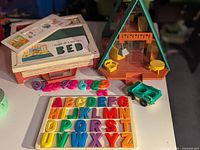 Vintage Fisher Price A-frame house with hanging light, chair, small round table; number pieces, alphabet letters on trays, small green wagon.