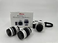 Pentax K2000 camera body with strap, two additional lenses, and original box arranged on white background