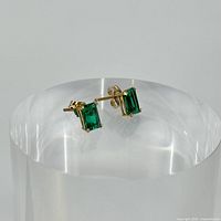 Pair of gold stud earrings with emerald green created stones shown from angled front view on clear cylindrical stand.