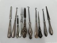 Top-down view of 10 assorted antique or vintage flatware pieces, including button hooks, forks, and knives, arranged side by side on white background