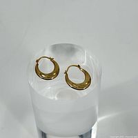 Pair of small 10k gold hoops with waved design placed on a clear round stand for display.