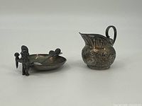 Photo of all three silver items: mini spoon with figure handle, petite ashtray with attached figure, and miniature ornate pitcher side by side.