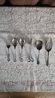 Full view of the six-piece antique silver-plated serving set including three spoons, one fork, tongs, and pie server arranged on lace tablecloth.