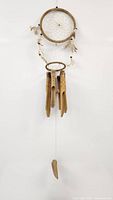 Full view of the feather dream catcher with web, feathers, and bamboo wind chimes hanging below against a white wall.
