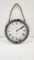 Front view of circular metal clock with distressed green finish and white Roman numeral face reading 'London 1883'. Thick twisted rope attached for hanging.
