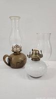 Two vintage oil lamps side by side on white background: pottery oil lamp with handle and clear chimney, white confetti glass oil lamp with burner and wick, separate clear chimney
