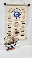 View of the sailor-themed fabric banner with various nautical knots and a blue ship wheel with 'Welcome Aboard' text, next to the detailed wooden sailboat model mounted on a base.
