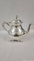 Side view of silver plated teapot showing curved handle, spout, lid with knob, and four feet.