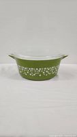 Side view of vintage avocado green Pyrex casserole dish with white daisy print and clear glass lid.
