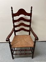 Full frontal photo of antique wooden arm chair with ladder back and rush seat against white wall.