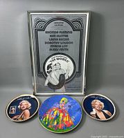 Photo showing the three Marilyn Monroe collectible plates arranged in front of the framed poster of 'The Women'. The poster is silver, black, and white with cast names listed.