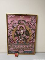 Framed mixed media painting of an Asian celestial goddess in a mandala setting with floral and cloud motifs, placed against a beige wall and shown with an apple for scale.