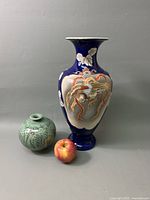 Two Asian vases with an apple for size reference. The taller cobalt blue vase features a detailed bird motif in relief and raised floral design. The smaller, round celadon vase has hand-painted floral patterns.