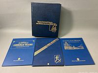 Four blue hardcover stamp folios displayed with gold embossed titles and designs including Norman Rockwell Classics and Golden Gate Bridge 50th Anniversary.