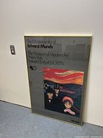 Framed gallery poster leaning against wall featuring Edvard Munch artwork and exhibition text