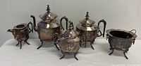 Group photo showing 2 teapots, 2 sugar bowls, and 1 creamer with ornate silverplate design, floral engravings, and decorative feet.