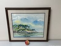 Framed watercolor painting showing coastal landscape with homes, trees, and water, in a wood frame with matting.