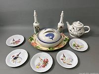 Full lot overview showing six dessert plates each with different gourmet food and wine prints, large floral platter underneath, covered casserole dish with lid featuring a blue fish, a floral patterned teapot with gold details, and two floral cruets with white stoppers.