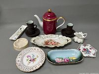 Full lot of assorted porcelain ware including teapot, creamers, decorative dishes and ashtray arranged on grey background.