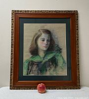 Full view of the framed pastel portrait with an apple for scale, showing the ornate gold outer frame and wooden inner frame with a dark green mat.