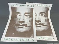 Five Salvador Dali posters laid flat showing large black and white face portrait with prominent mustache and printed text.
