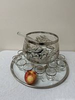 Full view of punch bowl with silver overlay, sixteen clear glass cups arranged around the base on a silver-rimmed tray, and an apple for size reference.